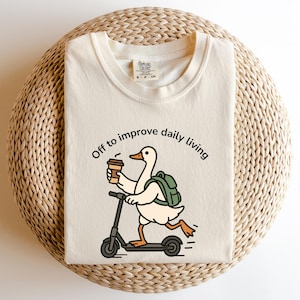 May include: Cream-colored t-shirt with a graphic of a duck riding a scooter, holding a coffee cup, and wearing a backpack. The text "Off to improve daily living" is printed above the duck. The shirt is folded on a woven, circular surface.