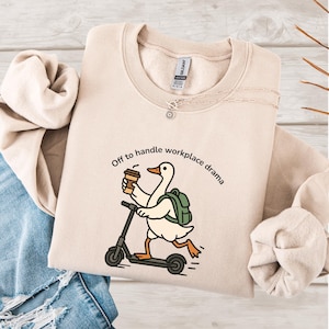 May include: Beige sweatshirt with a graphic of a goose riding a scooter, holding a coffee cup, and wearing a backpack. The text "Off to handle workplace drama" is printed above the goose. A silver necklace with a sun pendant is also visible.