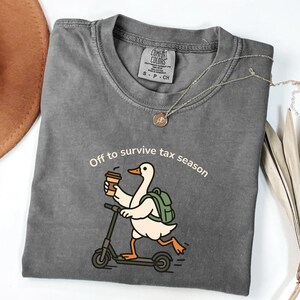 K&ouml;nnte beinhalten: Ein graues Comfort Colors T-Shirt mit dem Text "Off to survive tax season". Das Shirt zeigt eine Cartoon-Ente auf einem Roller, die eine Kaffeetasse h&auml;lt und einen Rucksack tr&auml;gt. Eine silberne Halskette mit Anh&auml;nger ist ebenfalls sichtbar.