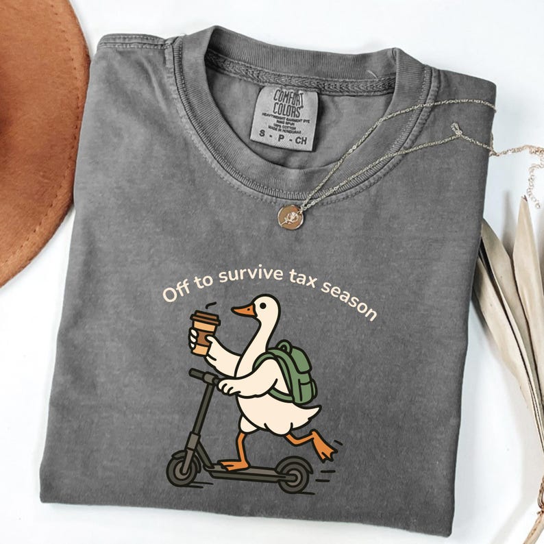 K&ouml;nnte beinhalten: Ein graues Comfort Colors T-Shirt mit dem Text "Off to survive tax season". Das Shirt zeigt eine Cartoon-Ente auf einem Roller, die eine Kaffeetasse h&auml;lt und einen Rucksack tr&auml;gt. Eine silberne Halskette mit Anh&auml;nger ist ebenfalls sichtbar.