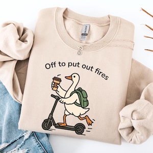 May include: A beige crewneck sweatshirt featuring a graphic of a white goose wearing a green backpack, riding a black scooter, and holding a coffee cup. The text above the goose reads, Off to put out fires, in black lettering.