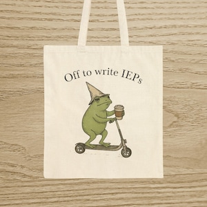 May include: Cream-coloured tote bag featuring a cartoon frog in a wizard hat, riding a scooter and holding a coffee cup. The text "Off to write IEPs" is printed above the frog illustration.