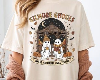 Może przedstawiać: Kremowa koszulka z grafiką o tematyce Halloween. Projekt zawiera dwie postacie duchów, altanę, dynie i tekst "GILMORE GHOULS WHERE YOU HAUNT, I WILL FOLLOW". Grafika jest w odcieniach brązu, pomarańczy i czerni.