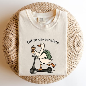 May include: A cream-coloured t-shirt featuring the phrase "Off to de-escalate" and a cartoon duck on a scooter, holding a coffee cup and wearing a backpack. The shirt is displayed on a woven, circular surface.