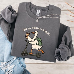 May include: A charcoal gray sweatshirt with the text "Off to adjust oxygen." The graphic shows a white goose riding a scooter, holding a coffee cup, and wearing a green backpack. A silver necklace with a sun pendant is also visible.