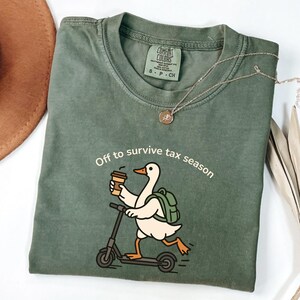 K&ouml;nnte beinhalten: Salbeigr&uuml;nes T-Shirt mit dem Schriftzug "Off to survive tax season" und einer Cartoon-Ente auf einem Roller, die eine Kaffeetasse h&auml;lt. Das Shirt hat ein Comfort Colors-Etikett und eine silberne Halskette.