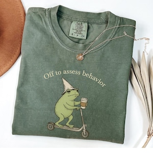 May include: Green Comfort Colors t-shirt with the phrase "Off to assess behavior" and a cartoon frog riding a scooter while holding a coffee cup. The shirt has a small tag at the neckline and a silver necklace.