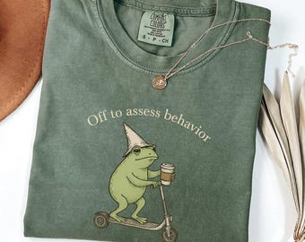May include: Green Comfort Colors t-shirt with the phrase "Off to assess behavior" and a cartoon frog riding a scooter while holding a coffee cup. The shirt has a small tag at the neckline and a silver necklace.