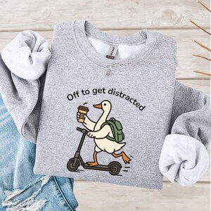 May include: A light grey sweatshirt with the slogan "Off to get distracted" printed in black. The graphic shows a cartoon goose on a scooter, holding a coffee cup and wearing a backpack.