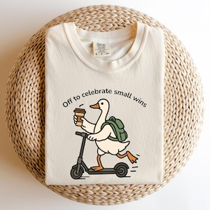 Könnte beinhalten: Cremefarbenes T-Shirt mit einer Grafik einer Ente auf einem Roller, die einen Kaffeebecher hält und einen Rucksack trägt. Der Text "Off to celebrate small wins" steht über der Ente. Das Shirt liegt auf einer gewebten, kreisförmigen Oberfläche.