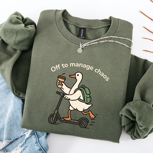 Puede incluir: Una sudadera de cuello redondo color verde oliva con un gráfico de un ganso blanco con pico y patas naranjas, que lleva una mochila verde, sostiene una taza de café y monta un patinete negro. El texto dice, Off to manage chaos.