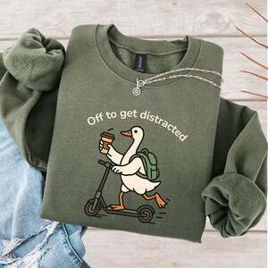 May include: An olive green sweatshirt featuring the text "Off to get distracted" and a cartoon goose on a scooter, holding a coffee. The goose has a backpack. A silver necklace is on the sweatshirt.