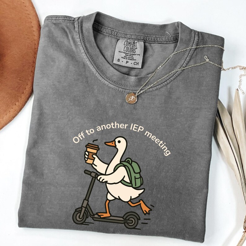 May include: Gray Comfort Colors t-shirt with the text "Off to another IEP meeting." A cartoon duck with a backpack and coffee cup rides a scooter. A silver necklace with a pendant is also visible.