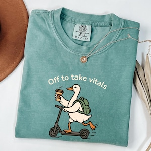 May include: Teal Comfort Colors t-shirt with a graphic of a goose riding a scooter, holding a coffee cup, and wearing a backpack. The text "Off to take vitals" is above the goose. A silver necklace with a pendant is on the shirt.