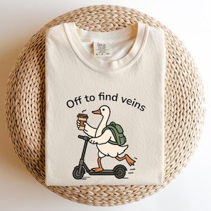 Op de afbeelding: Crèmekleurig T-shirt met de tekst "Off to find veins" en een cartoon eend op een scooter, met een koffiekopje en een rugzak. Het shirt is opgevouwen op een geweven, cirkelvormig oppervlak.