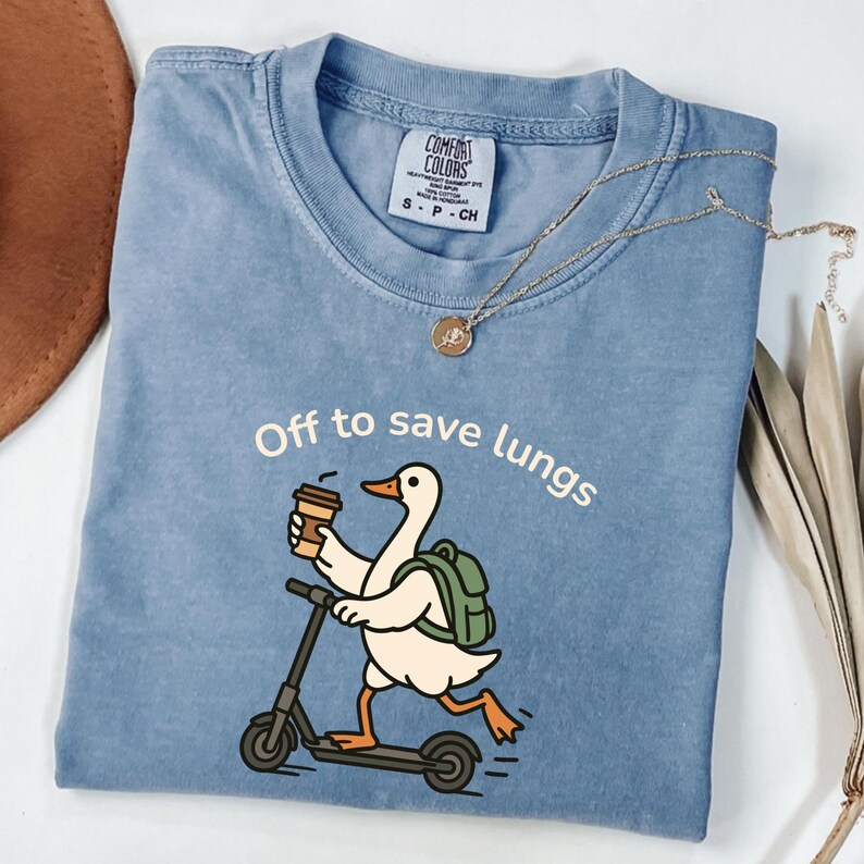 May include: A blue Comfort Colors t-shirt with the phrase "Off to save lungs" and a cartoon duck riding a scooter while holding a coffee cup. The duck has a green backpack. A gold necklace is draped on the shirt.