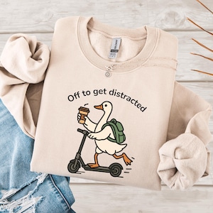 May include: A beige sweatshirt featuring the text "Off to get distracted" and a cartoon goose on a scooter, holding a coffee cup and wearing a backpack. The sweatshirt is folded on a light wood surface.