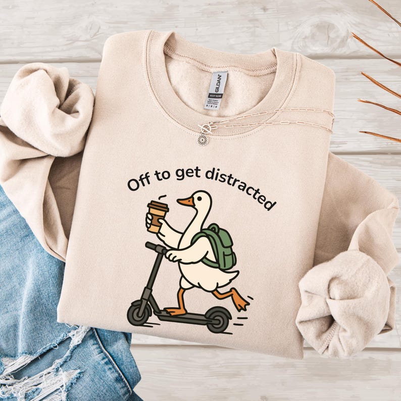 May include: A beige sweatshirt featuring the text "Off to get distracted" and a cartoon goose on a scooter, holding a coffee cup and wearing a backpack. The sweatshirt is folded on a light wood surface.