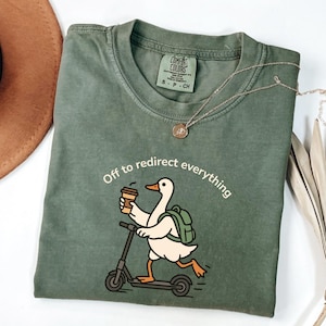 May include: Green Comfort Colors t-shirt with the phrase "Off to redirect everything." The shirt features a cartoon duck riding a scooter while holding a coffee cup and wearing a backpack. A silver necklace is draped on the shirt.