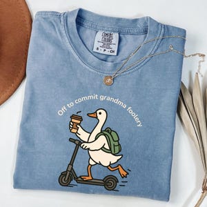 Puede incluir: Camiseta azul Comfort Colors con la frase "Off to commit grandma foolery" y un dibujo de un pato en un scooter con una taza de café y una mochila verde. Un collar dorado está sobre la camiseta.