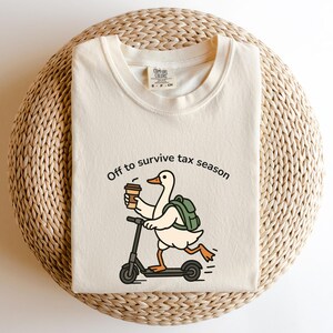 K&ouml;nnte beinhalten: Cremefarbenes T-Shirt mit dem Text "Off to survive tax season". Die Grafik zeigt eine Cartoon-Ente, die mit einem Roller f&auml;hrt, einen Kaffeebecher h&auml;lt und einen Rucksack tr&auml;gt. Das Shirt wird auf einer gewebten Oberfl&auml;che pr&auml;sentiert.