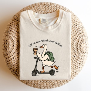 May include: Cream-colored t-shirt with the phrase "Off to overthink everything." The graphic features a cartoon duck riding a scooter while holding a coffee cup and wearing a backpack. The shirt is on a woven surface.