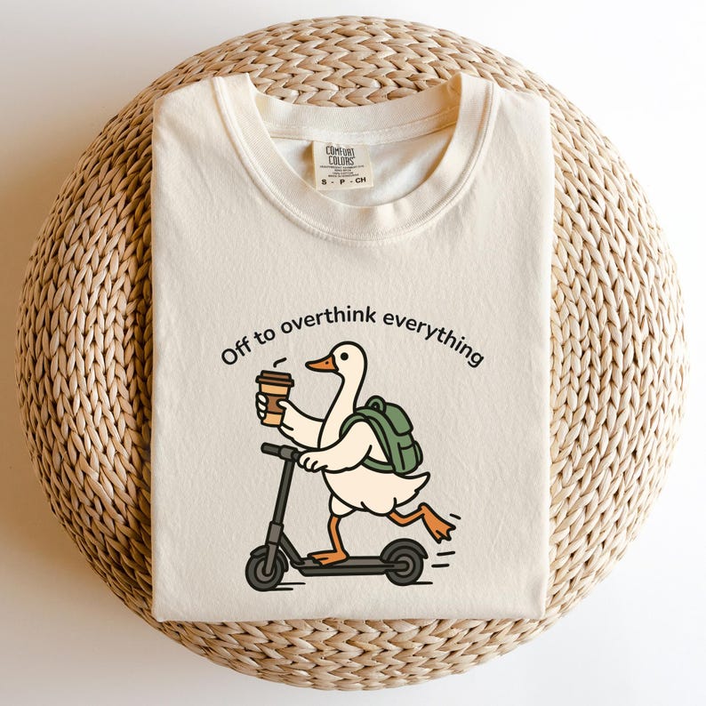 May include: Cream-colored t-shirt with the phrase "Off to overthink everything." The graphic features a cartoon duck riding a scooter while holding a coffee cup and wearing a backpack. The shirt is on a woven surface.