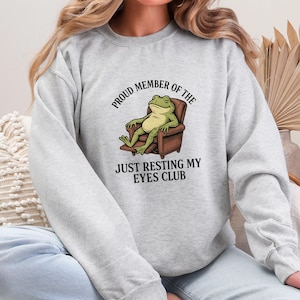 May include: A light gray sweatshirt featuring a relaxed frog illustration in a brown armchair. The text reads "PROUD MEMBER OF THE JUST RESTING MY EYES CLUB". The frog is green and the armchair is brown. The sweatshirt is a casual style.