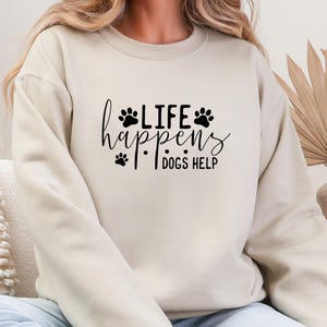 May include: A beige sweatshirt with the text "Life Happens Dogs Help" in black script and paw print graphics. The sweatshirt is a neutral color and has a relaxed fit, suitable for casual wear.