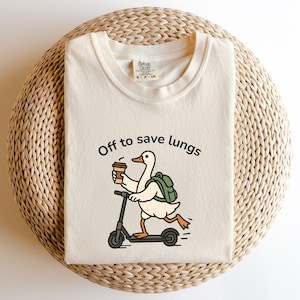 May include: Cream-colored t-shirt with the text "Off to save lungs." The graphic shows a cartoon goose riding a scooter while holding a coffee cup and wearing a backpack. The shirt is made of a soft material.