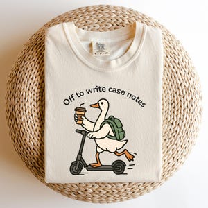 May include: Cream-colored t-shirt with the text "Off to write case notes." A cartoon goose with a backpack and coffee cup rides a scooter. The shirt is laid on a woven, circular surface.