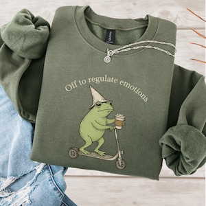 May include: An olive green sweatshirt with the text "Off to regulate emotions". The graphic shows a green frog on a scooter with a coffee cup. A silver necklace is draped on the sweatshirt. The sweatshirt is folded on a wooden surface.