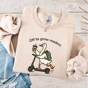 Może przedstawiać: Beżowa bluza z napisem "Off to grow readers" i kreskówkową gęsią na skuterze trzymającą filiżankę kawy. Gęś ma zielony plecak. Srebrny naszyjnik z zawieszką w kształcie słońca znajduje się na bluzie.