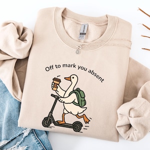 May include: A beige crewneck sweatshirt featuring a graphic of a white goose with an orange beak and feet, wearing a green backpack and holding a brown coffee cup while riding a black scooter. The text above the goose reads, Off to mark you absent.