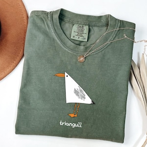 May include: An olive green crew neck t-shirt featuring a graphic of a white triangular seagull with an orange beak and feet. The bird has a grey wing patch and the text "triangull" printed below it. The shirt has a Comfort Colors brand label.
