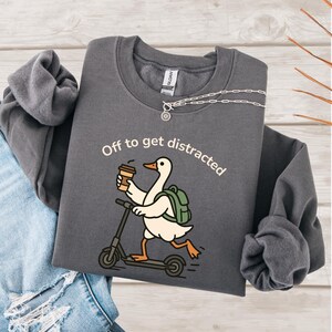 May include: Charcoal grey sweatshirt featuring the phrase "Off to get distracted." The design depicts a cartoon goose on a scooter, carrying a coffee cup and a backpack. A silver necklace with a sun pendant is also visible.