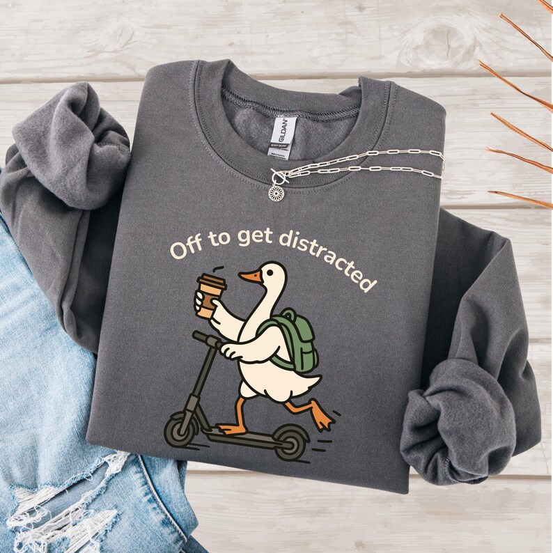 May include: Charcoal grey sweatshirt featuring the phrase "Off to get distracted." The design depicts a cartoon goose on a scooter, carrying a coffee cup and a backpack. A silver necklace with a sun pendant is also visible.