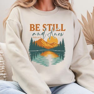 Może przedstawiać: Beżowa bluza z nadrukiem krajobrazu. Grafika przedstawia pasmo górskie, słońce, drzewa i wodę z napisem "BE STILL and KNOW" i "Psalm 46:10". Projekt jest w odcieniach pomarańczu, turkusu i beżu.