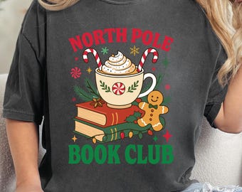 Puede incluir: Camiseta gris oscuro con un diseño festivo. El gráfico incluye una taza de chocolate caliente con crema batida, bastones de caramelo, un hombre de jengibre y las palabras "NORTH POLE BOOK CLUB" en rojo y verde.