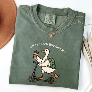 Op de afbeelding: Olijfgroen t-shirt met de tekst "Off to teach tiny humans". Een cartoon eend met een rugzak en koffiekopje rijdt op een scooter. Een zilveren ketting met een kleine hanger ligt op het shirt.