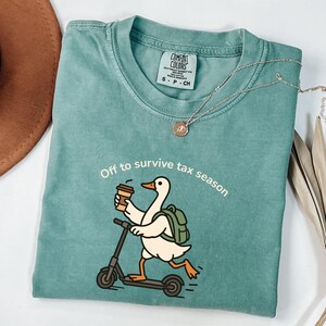 K&ouml;nnte beinhalten: T&uuml;rkisfarbenes Comfort Colors T-Shirt mit einer Grafik einer Ente auf einem Roller, die Kaffee h&auml;lt und einen Rucksack tr&auml;gt. Der Text lautet "Off to survive tax season". Eine silberne Halskette mit Anh&auml;nger befindet sich auf dem Shirt.