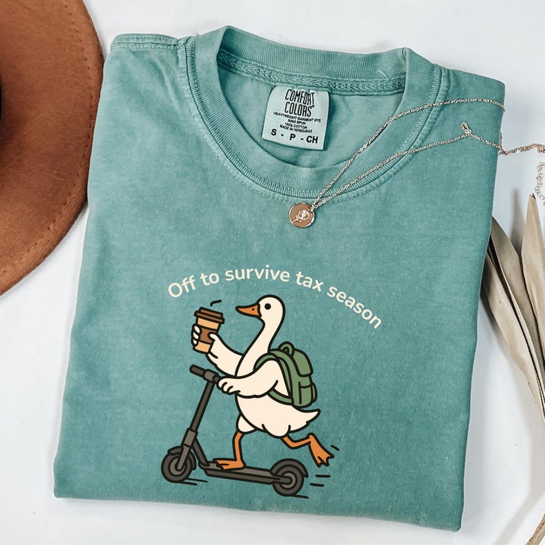 K&ouml;nnte beinhalten: T&uuml;rkisfarbenes Comfort Colors T-Shirt mit einer Grafik einer Ente auf einem Roller, die Kaffee h&auml;lt und einen Rucksack tr&auml;gt. Der Text lautet "Off to survive tax season". Eine silberne Halskette mit Anh&auml;nger befindet sich auf dem Shirt.