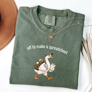 May include: An olive green t-shirt with a graphic of a white goose wearing a green cap and glasses, carrying a backpack and a clipboard. The text "off to make a spreadsheet" is printed in white above the goose. The shirt has a Comfort Colors label.