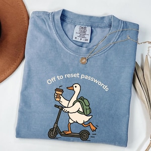 May include: A blue t-shirt featuring a graphic of a white goose with an orange beak and feet, wearing a green backpack, riding a black scooter, and holding a coffee cup. The text above the goose reads, Off to reset passwords in white font.