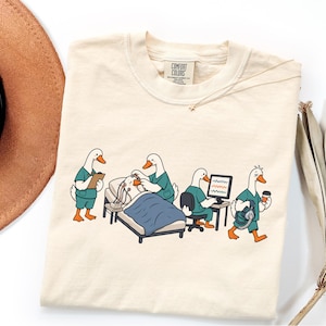 May include: A cream-colored t-shirt featuring a graphic of four white ducks dressed in teal medical scrubs. One duck holds a clipboard, another attends to a patient in a bed, one sits at a computer monitor, and the last carries medical equipment.