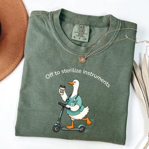 Puede incluir: Una camiseta verde oliva con el gráfico de un ganso blanco que viste una casaca médica color verde azulado y un estetoscopio, montado en un patinete mientras sostiene un café. El texto sobre el ganso dice, Off to sterilize instruments.