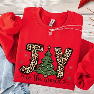 May include: A red sweatshirt with the word "JOY" in leopard print and a green Christmas tree graphic. The phrase "to the world" is written below the tree. The sweatshirt has a crew neck and long sleeves.