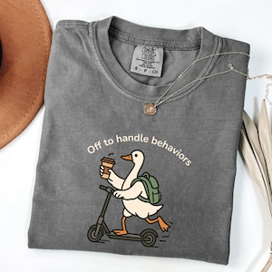 May include: A grey Comfort Colors t-shirt featuring the text "Off to handle behaviours" and a cartoon duck on a scooter, holding a coffee cup. The duck has a green backpack. A silver necklace and a brown hat are also visible.