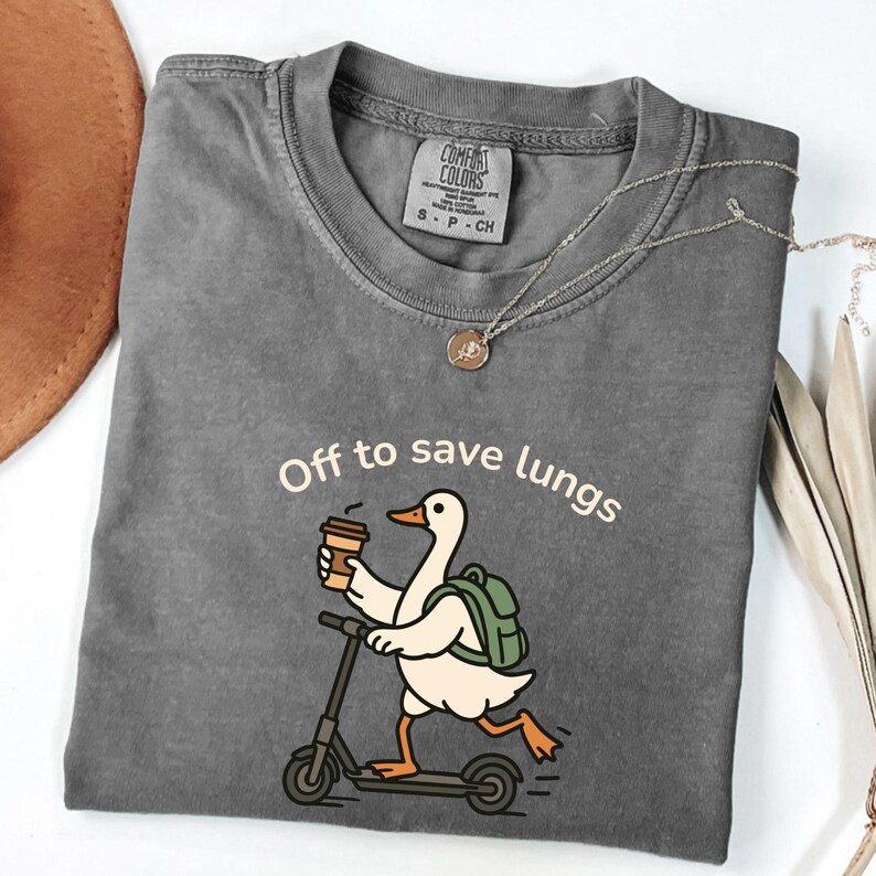May include: Gray Comfort Colors t-shirt with the phrase "Off to save lungs." The graphic shows a white duck riding a scooter while holding a coffee cup and wearing a backpack. A silver necklace with a pendant is also visible.