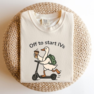 May include: Cream-colored t-shirt with the text "Off to start IVs." The graphic shows a cartoon duck riding a scooter while holding a coffee cup and wearing a backpack. The shirt is folded on a woven, circular surface.